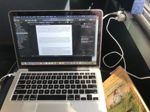 Amtrak writing set-up. Laptop on the left and iPhone charging cord both plugged into an outlet under the window to the right, with research book to the right.
