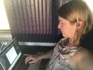 Profile picture of myself, with laptop on lap and corded earbud in ear. Curtain shading the window in the background. Laptop and iPhone charging cords both plugged into outlet beneath the window.