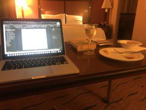 Writing set-up at the Homewood Suites in Fort Worth. Laptop on the left, research books and glass of wine in the middle, mostly-eaten plate and bowl of dinner on the right. Bed in the background. Soft yellow lighting, indicating picture was taken after dark.